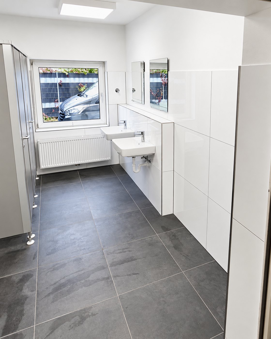 Sanierter Sanitärraum mit großformatigen dunklen Bodenfliesen, weißen Wandfliesen und Waschtischzeile